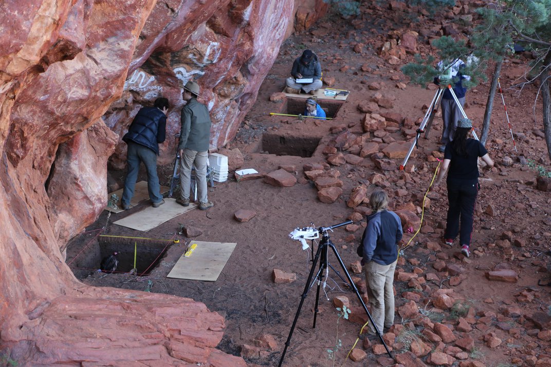 Un equipo de arqueólogos realiza trabajo de campo, excava agujeros cuadrados y usa microscopios en tierra de color rojo oscuro.
